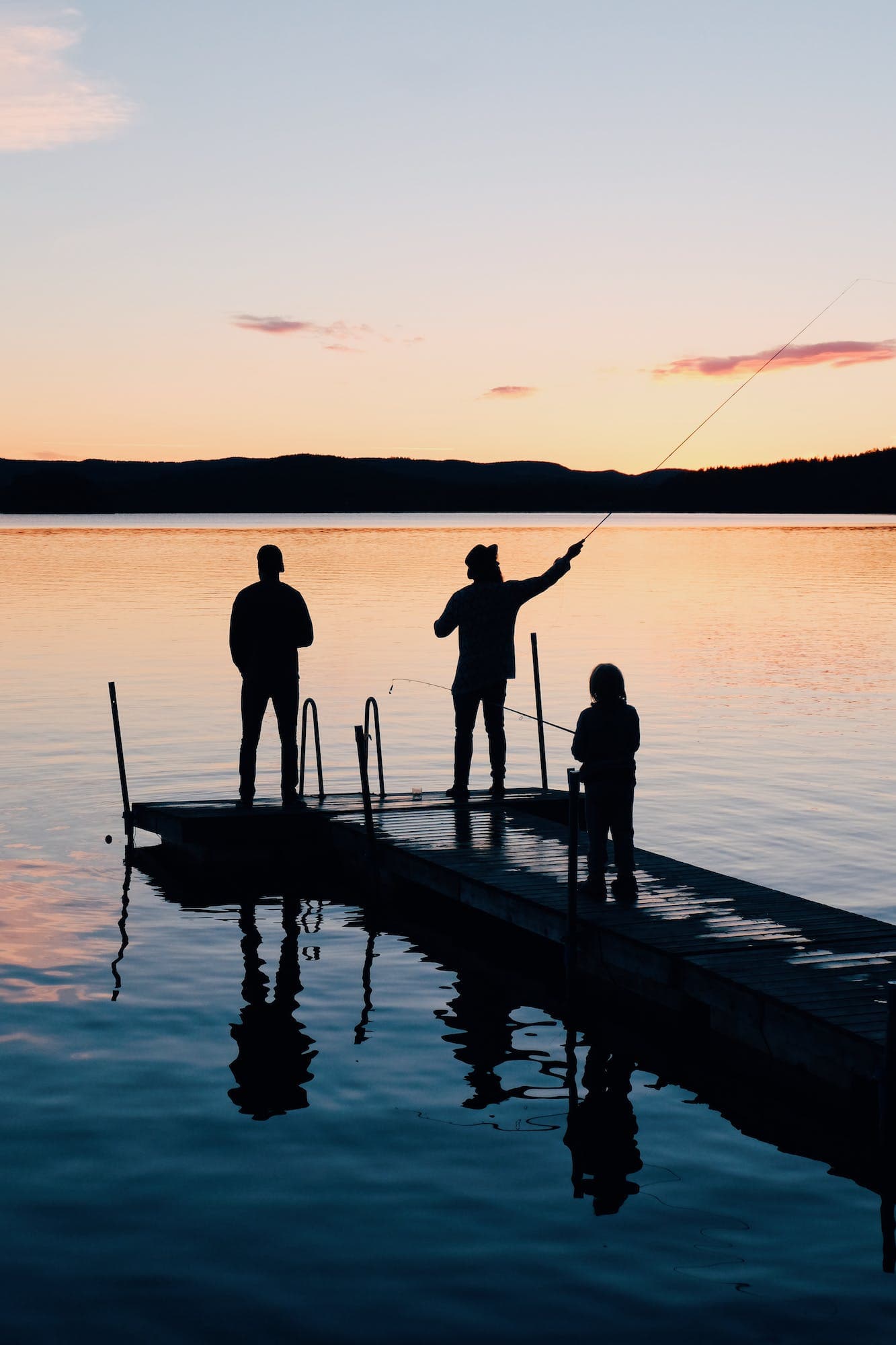 Kolme eri-ikäistä henkilöä kalastamassa laiturilla. Kuva on otettu auringonlaskun aikaan ja henkilöt ovat silhuetteja.