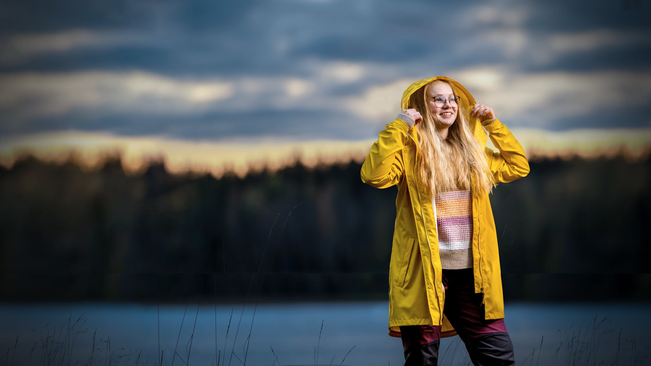 Nuori nainen, jolla pitkät vaaleat hiukset seisoo illalla järven rannalla keltainen sadetakki päällään.