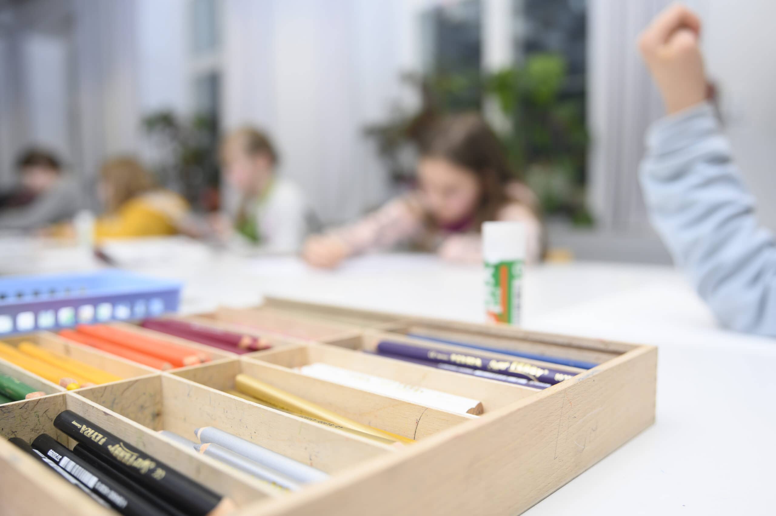 Kuvassa etualalla laatikko jossa erivärisiä kyniä ja taustalla sumeana lapsia askartelemassa ja piirtämässä.