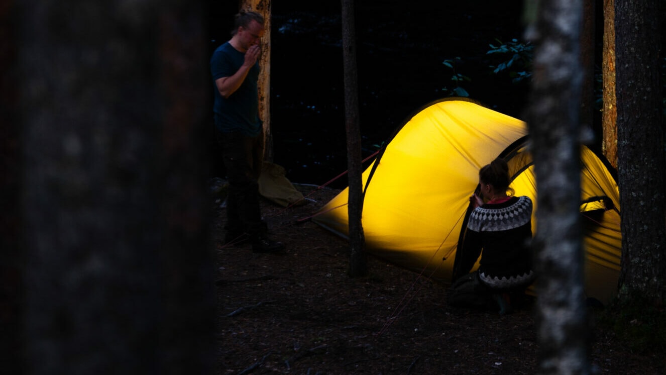 Kaksi henkilöä telttailemassa pimeässä. Toinen henkilö seisoo ja toinen istuu keltaisen teltan edessä.